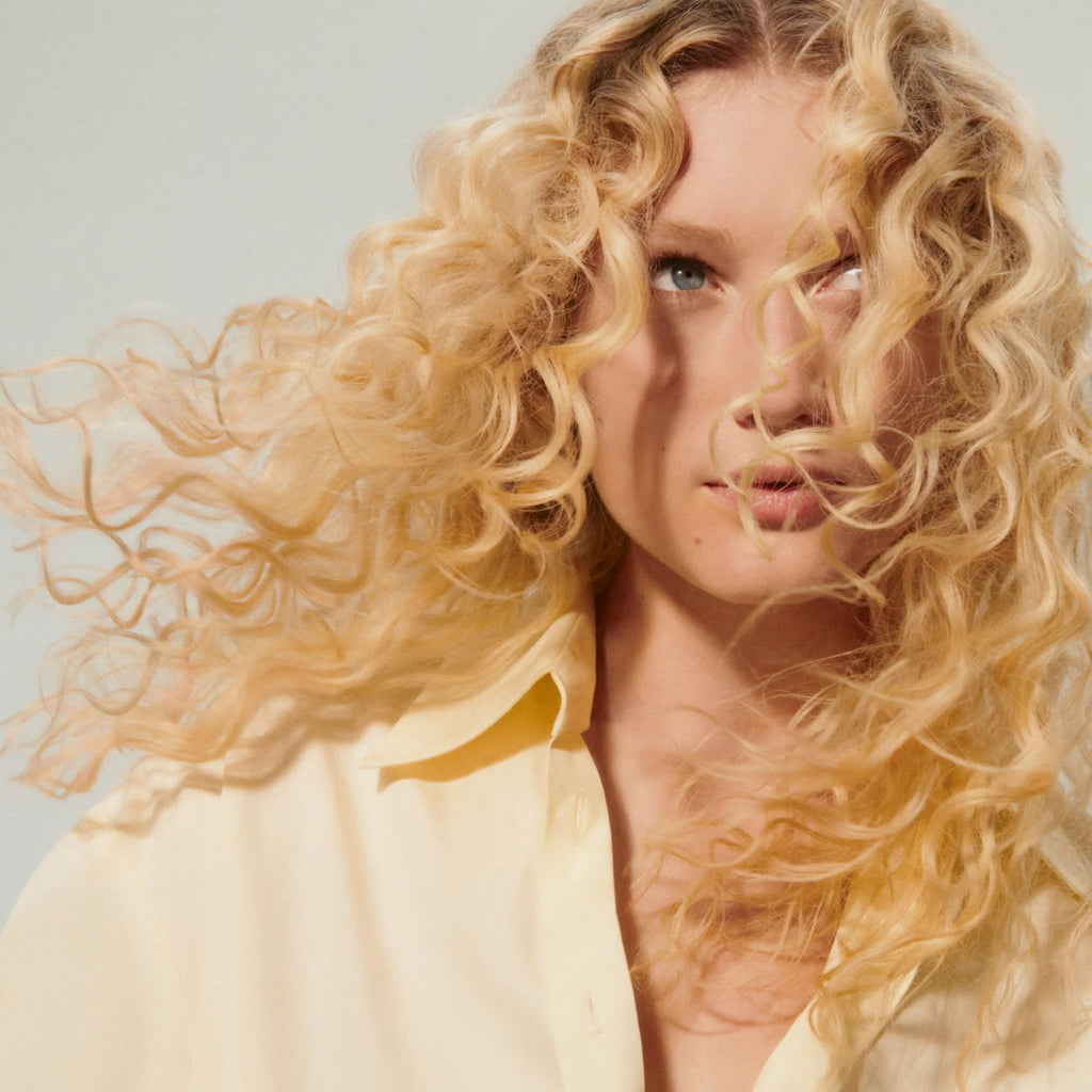Woman with blonde curly hair wearing a light-colored top against a neutral background