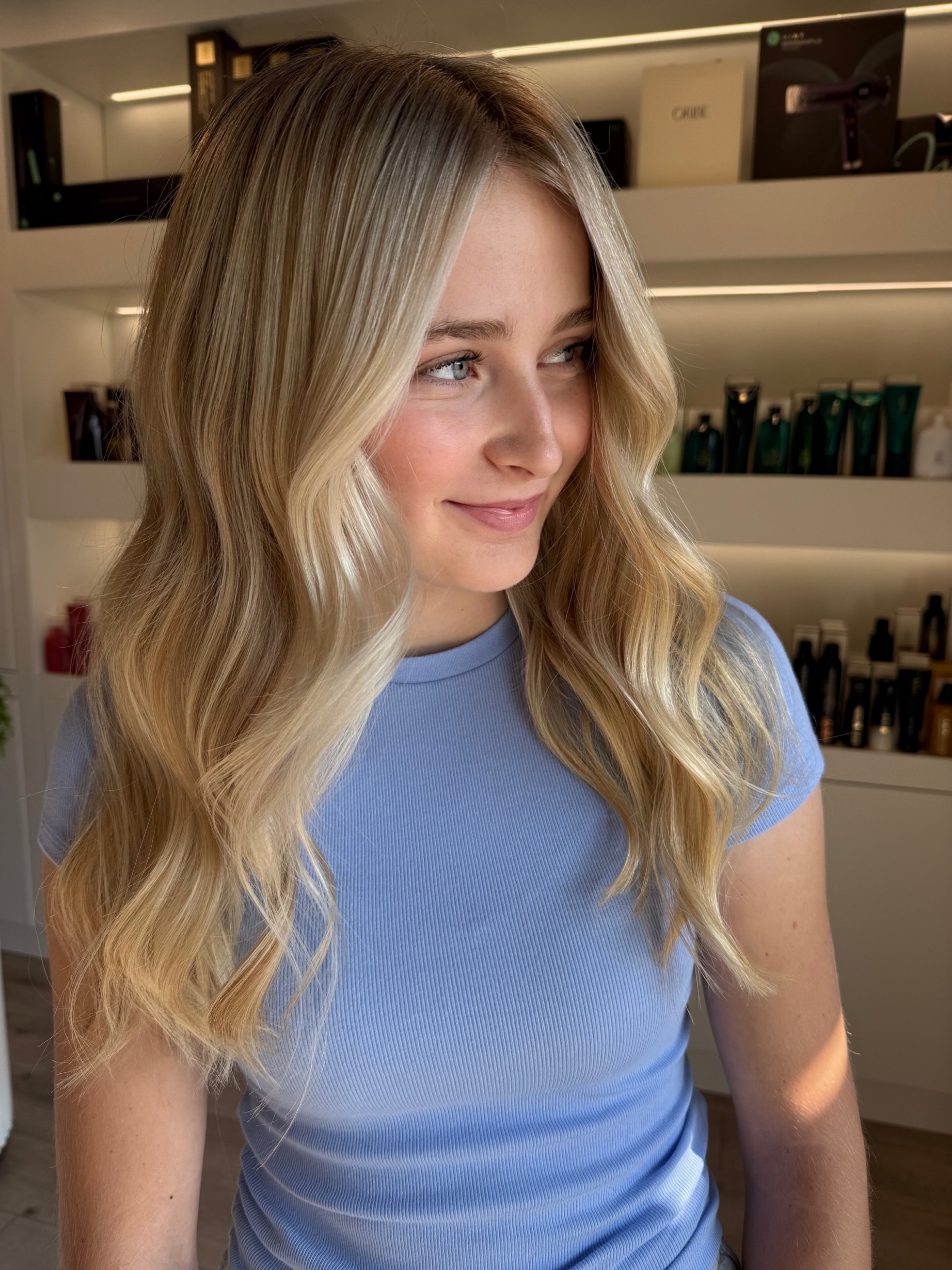 Woman with blonde hair wearing a blue top in a room with shelves in the background